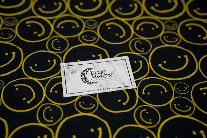 Flat sample shot of All Smiles Printed Spandex with a Blue Moon Fabrics standard size business card laid on top of the print for pattern sizing. The print is of vibrant yellow smiley faces of various sizes on a black background.