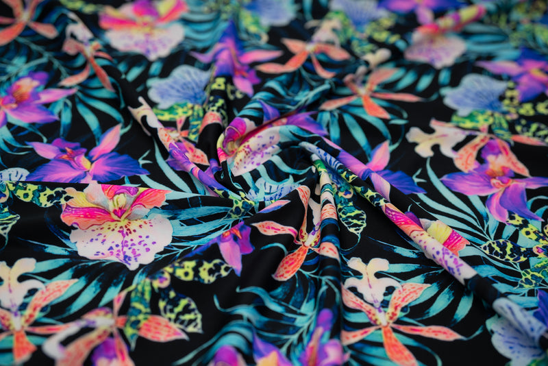 Swirled sample of Irises and Palm Fronds Printed Spandex Fabric. The print is of vibrant colored Irises and other exotic flowers over bright light blue fronds on a black background.