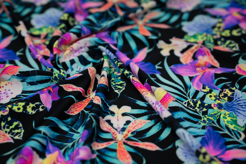 Swirled sample of Irises and Palm Fronds Printed Spandex Fabric. The print is of vibrant colored Irises and other exotic flowers over bright light blue fronds on a black background.