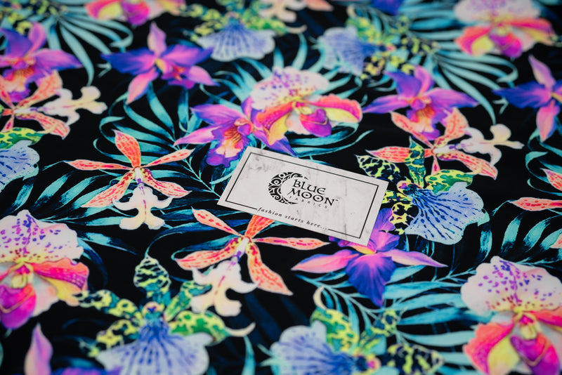 Flat sample of Irises and Palm Fronds Printed Spandex Fabric with a Blue Moon Fabrics standard size business card laid on top of the print for pattern scale. The print is of vibrant colored Irises and other exotic flowers over bright light blue fronds on a black background.