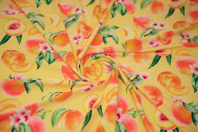 Swirled sample shot of It's All Peachy Printed Spandex Fabric. The print is of whole peach clusters with pink peach blossoms and green leaves, whole individual peaches with peach slices and peach halves on a yellow toned background.  