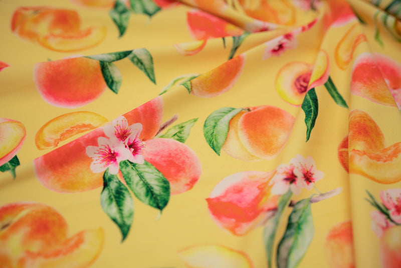 Swirled sample shot of It's All Peachy Printed Spandex Fabric. The print is of whole peach clusters with pink peach blossoms and green leaves, whole individual peaches with peach slices and peach halves on a yellow toned background.  