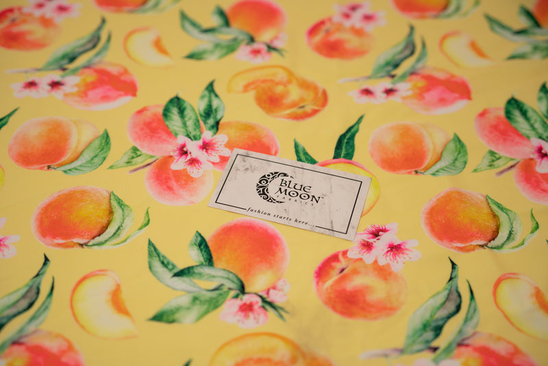Flat sample shot of It's All Peachy Printed Spandex Fabric with Blue Moon Fabrics standard size business card laid on top of print for scale perspective. The print is of whole peach clusters with pink peach blossoms and green leaves, whole individual peaches with peach slices and peach halves on a yellow toned background.  