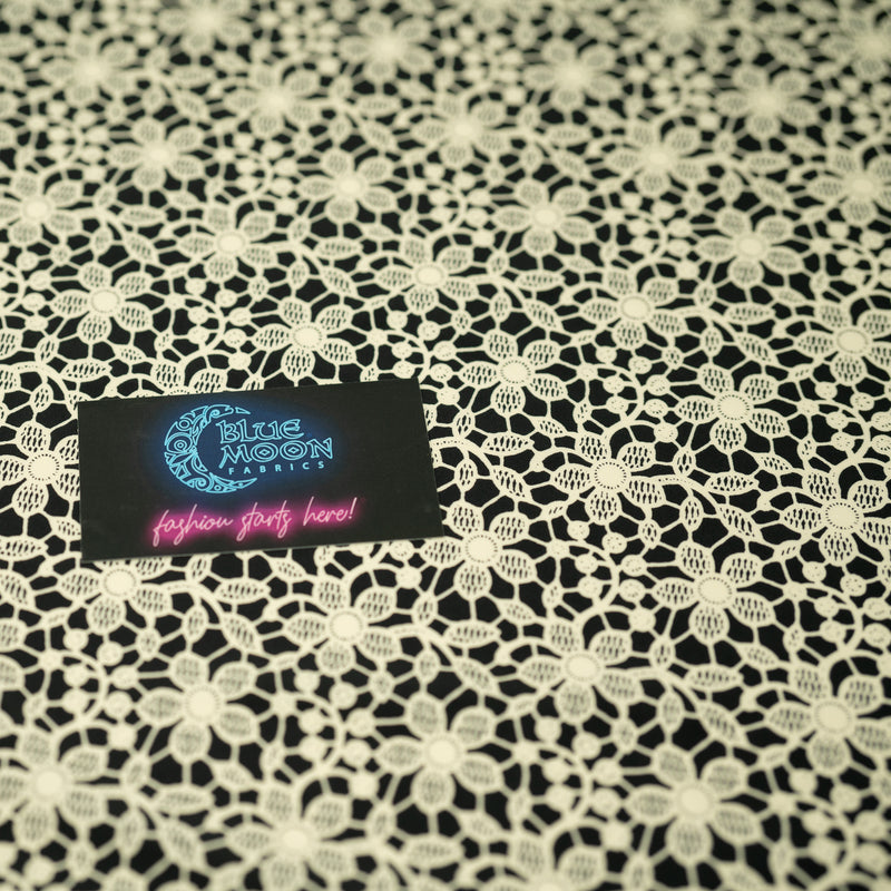 A flat sample of Ivory Flower Lace Pattern on Black Printed Spandex with a Blue Moon Fabrics standard size business card laid on top of the print for pattern sizing.