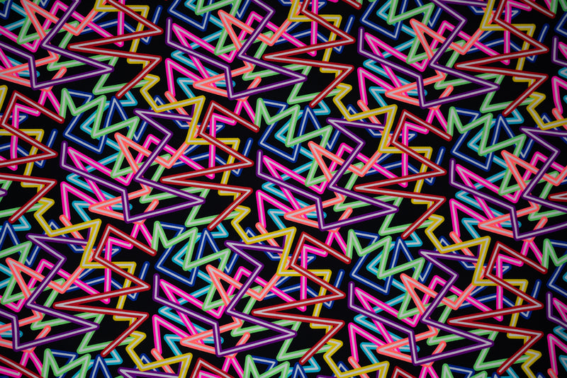 Flat sample shot of Retro Neon Lights Printed Spandex Fabric. The print is of zig-zag neon strobes in various colors of red, yellow, neon pink, light green, hot coral and blue with white as the filling on a black background.