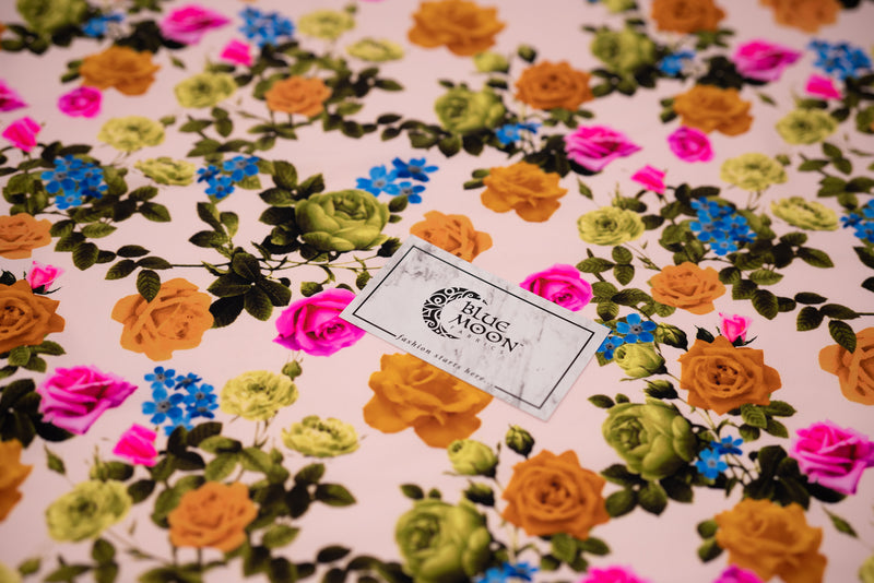 Detailed shot of Rose Garden Printed Spandex Fabric with Blue Moon Fabrics standard size business card laid on top for scale perspective. The print is of fluorescent pink, olive green and burnt orange roses on olive green colored stems with leaves and tiny blue flowers on a cream pink background. 