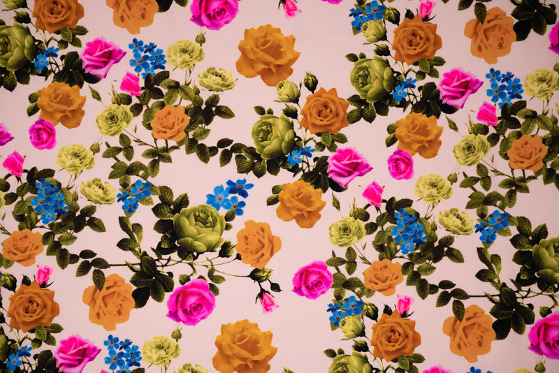Detailed shot of a flat piece of Rose Garden Printed Spandex Fabric with Blue Moon Fabrics standard size business card laid on top for scale perspective. The print is of fluorescent pink, olive green and burnt orange roses on olive green colored stems with leaves and tiny blue flowers on a cream pink background.