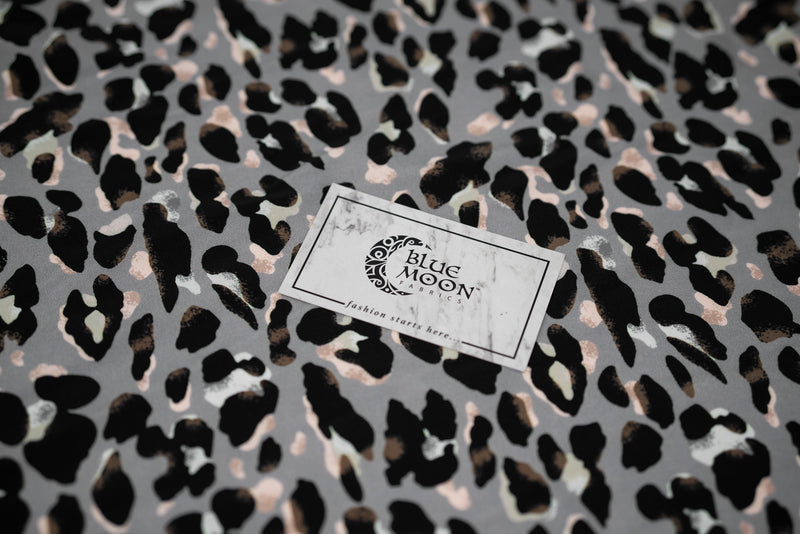 Flat sample shot of Sporty Leopard Printed Spandex Fabric with a Blue Moon Fabrics standard size business card laid on top for scale of pattern perspective. The print is of black leopard spots with light dust brown accents and light colored shadows on a gray background.